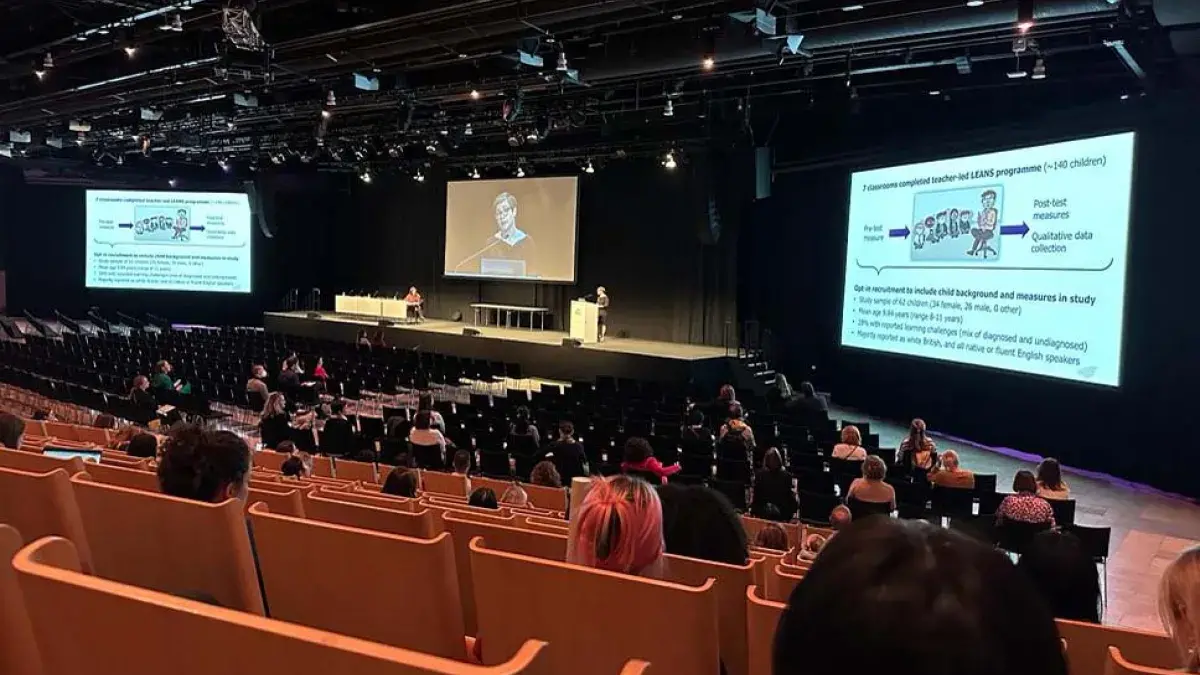 A lecture theatre full of people watching Dr Alyssa Alcorn give a talk. A video of Alyssa is projected on a big screen in the middle and two other big screens show her slides.