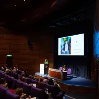 Photo of lecture theatre with speaker at lecturn in front of digital screen and audience watching
