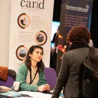 Photo of woman sitting at table in front of poster entitled 'Carid' and chatting with a standing person