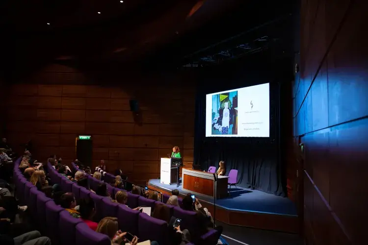 Photo of lecture theatre with speaker at lecturn in front of digital screen and audience watching