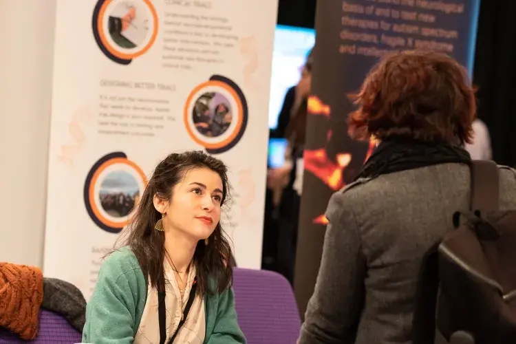 Photo of woman sitting at table in front of poster entitled 'Carid' and chatting with a standing person