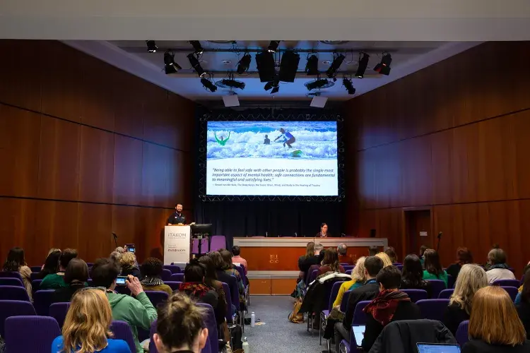 Photo of lecture theatre with a speaker, behind which is a screen showing a picture of someone sea surfing 